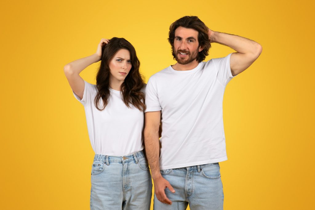 Perplexed young couple in white t-shirts and jeans, scratching their heads with puzzled expressions